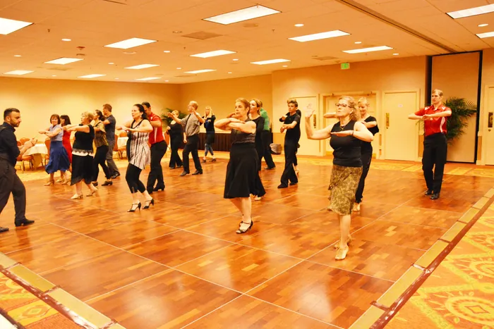 Students in loose rows on the ballroom floor, arms crossed at chest height, working through a figure together
