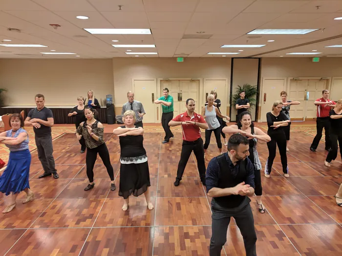 A room of students practising with arms crossed in front, an instructor leading from the front of the frame