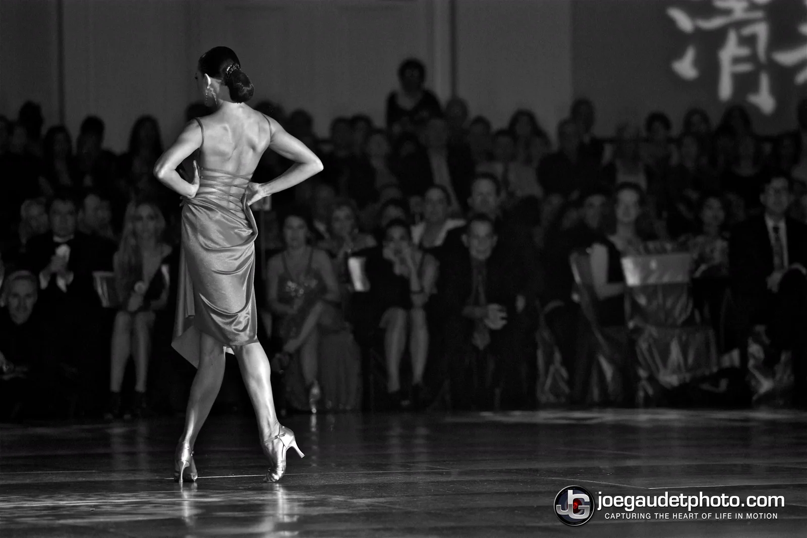 A dancer standing alone on the floor with her back to the camera, facing a packed audience