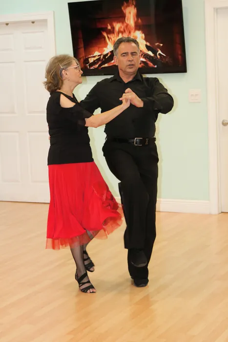 Lourdes in a red skirt and Ivan moving together past the fireplace screen