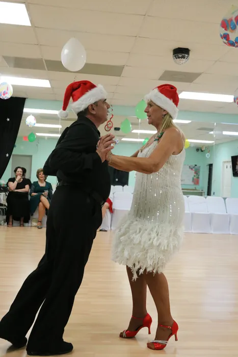 Peggie in a white feather dress and Ivan, both wearing Santa hats, dancing in a closed hold