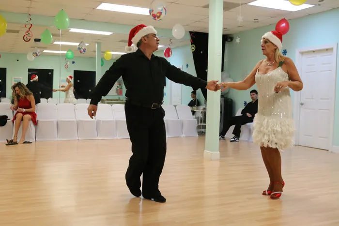 Peggie and Ivan in Santa hats, arms extended apart mid-step across the studio floor